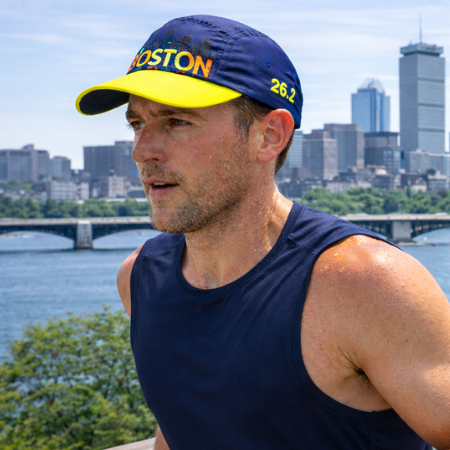 A man outdoors wearing a navy blue Boston 26.2 cap with a yellow brim and a navy sleeveless athletic tank top, with a city skyline and river in the background.