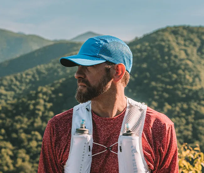 Man wearing a blue crusher hat and red shirt with a white backpack in a mountainous landscape