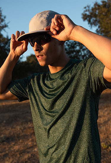A man outdoors at sunset wearing a dark green heathered athletic t-shirt, a beige performance running hat from Headsweats, and black sunglasses, adjusting his hat with both hands.