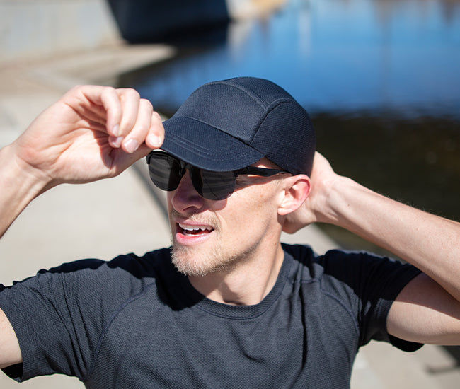 A man outdoors wearing a black performance running hat from Headsweats, black sunglasses, and a dark gray athletic t-shirt, with his hands adjusting the brim and back of the cap.