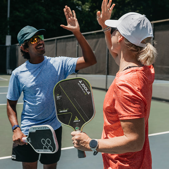 Two people on a pickleball court high-fiving, each holding a Joola paddle. The person on the left wears a blue t-shirt, black shorts, a green Headsweats pickeball hat, and gold-tinted sunglasses, while the person on the right wears an orange t-shirt, a white Headsweats pickleball cap, and a watch.